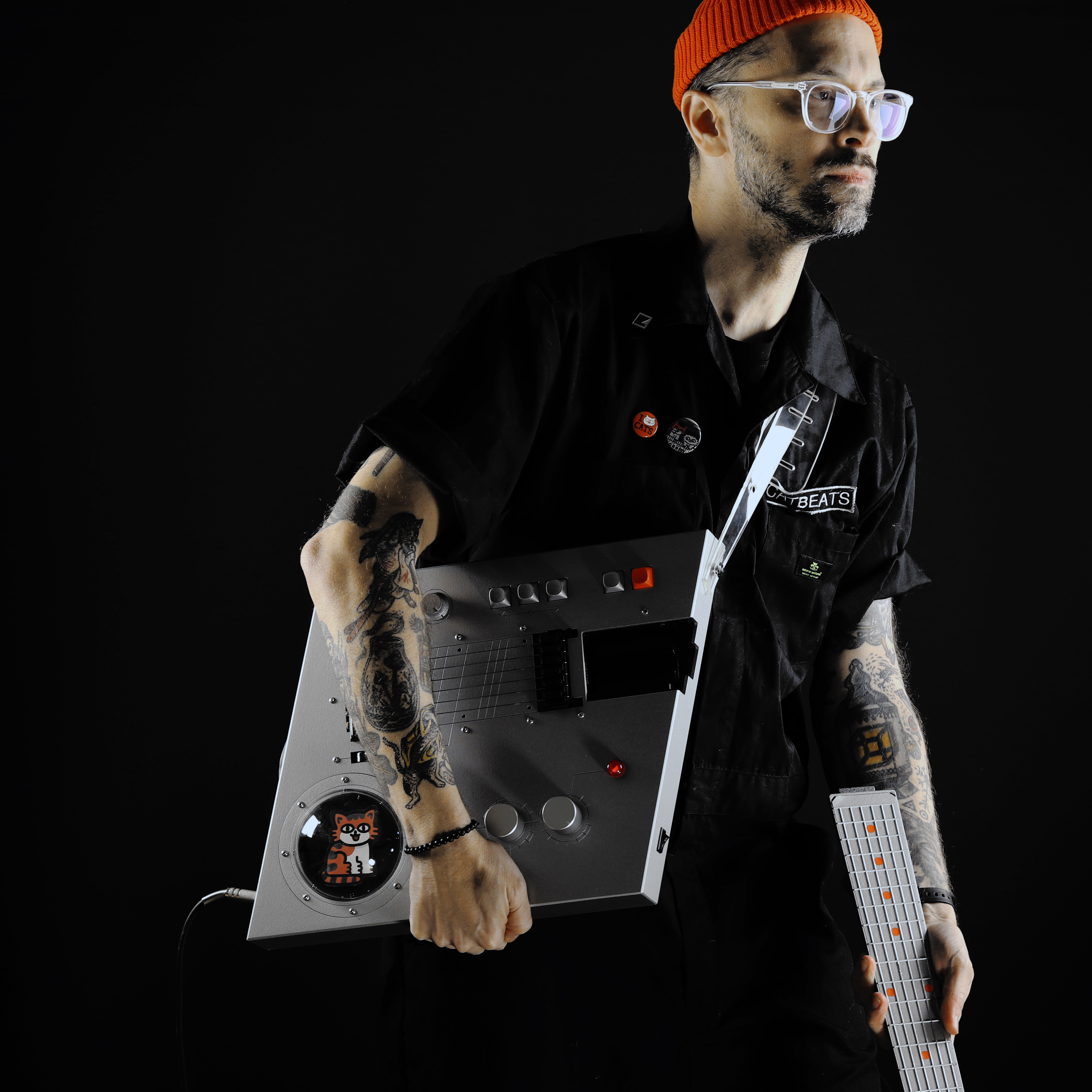 Orange hatted bespeckled designer holding rhomboid guitar body and wearing a black jumpsuit