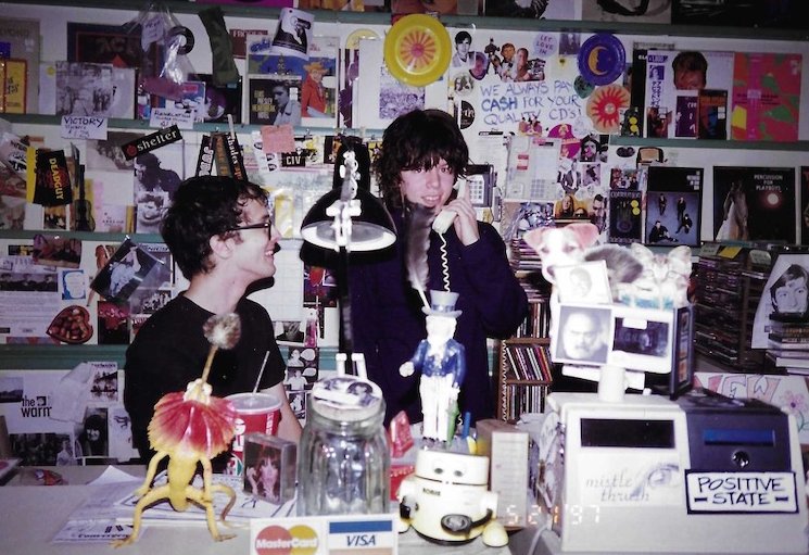 Two clerks behind the counter at Record Convergence, walls covered in show flyers and posters